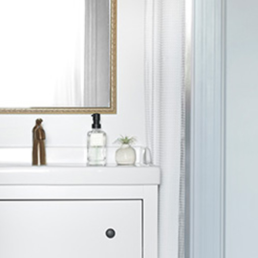 A porcelain sink with white cabinets and bronze hardware. A soap dispenser and a small succulent sit on top of the sink, along with a bronze-framed mirror above the sink. To the right of the sink is a curtain to for the shower and a light-blue painted wall.