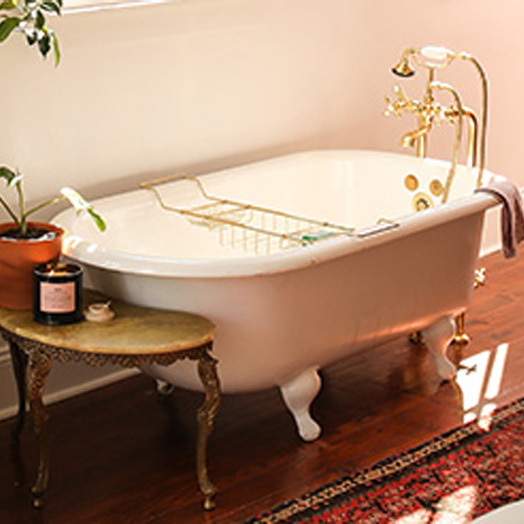 A bathtub with gold hardware. A side table is at the end of the bathtub with a candle and plant sitting on top of it.
